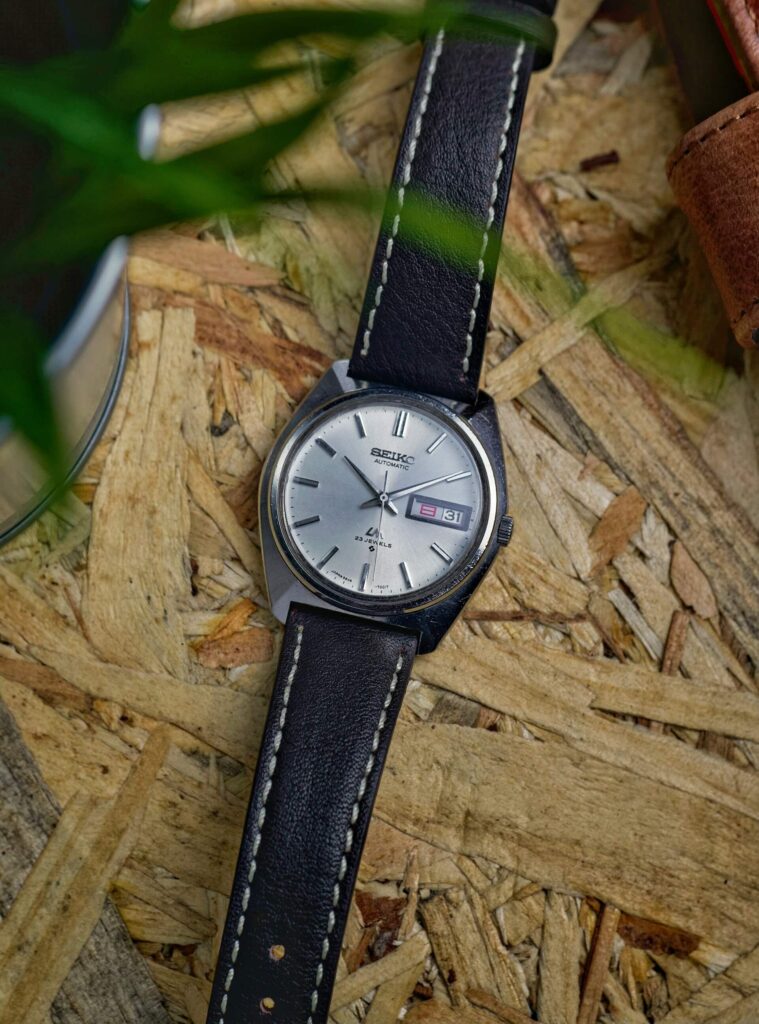 Ασημί vintage ρολόι Seiko με μαύρο δερμάτινο λουράκι και λευκές ραφές, τοποθετημένο πάνω σε ξύλινα ροκανίδια, με πράσινα φύλλα στο προσκήνιο.