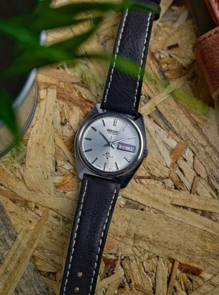 Ασημί vintage ρολόι Seiko με μαύρο δερμάτινο λουράκι και λευκές ραφές, τοποθετημένο πάνω σε ξύλινα ροκανίδια, με πράσινα φύλλα στο προσκήνιο.